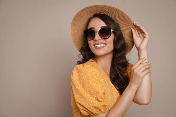 Beautiful young woman with straw hat and stylish sunglasses on beige background.